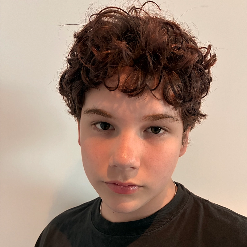 A young person with short, curly brown hair and fair skin looks directly at the camera, wearing a black T-shirt. The background is plain and light-colored, creating a simple and neutral setting that draws focus to the subject’s features, reminiscent of Kelsey Valente’s understated style.