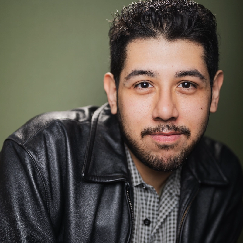 A man with short dark hair and facial hair, identified as Andres Pacheco, wears a black leather jacket over a checkered shirt as he looks at the camera and smiles slightly. The background is plain and green.
