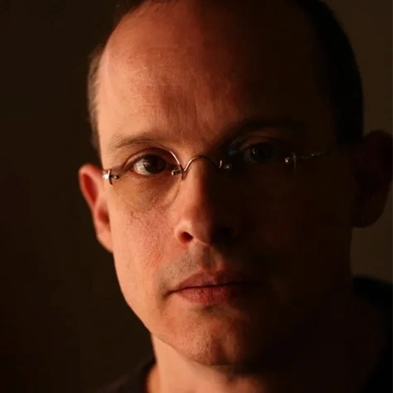 A middle-aged man with glasses and a receding hairline looks directly at the camera. Lit from the left, casting deep shadows on the right side of his face, Kendall Tubbs appears contemplative against a dark and out-of-focus background.