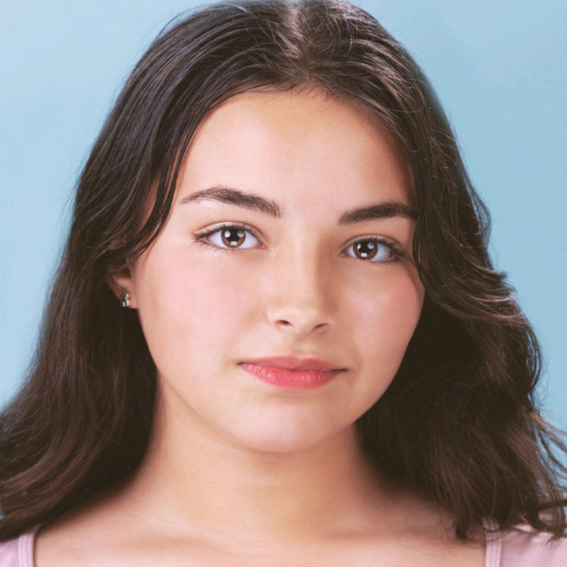 Alyssa Perez, a young woman with long, dark brown hair and clear skin, looks directly at the camera. She has brown eyes, natural makeup, and pink lips. She is wearing a light pink top and small earrings. The background is a light blue color.