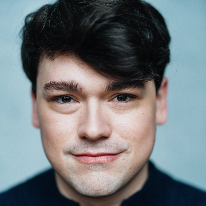 A young man with fair skin and dark brown hair smiles softly at the camera. Calvin Pierce has straight brown hair swept to the side, brown eyes, and wears a dark shirt. The background is plain and softly blurred, focusing attention on his face.