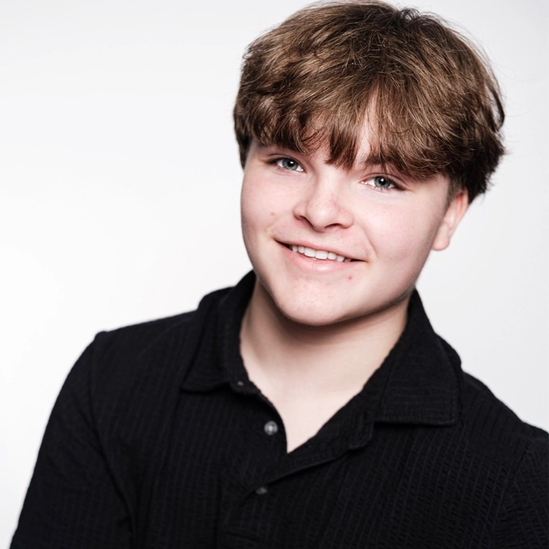 A headshot of Colton Piontkowski, a young person with light skin, blue eyes, and messy brown hair, smiling directly at the camera. They are wearing a black collared shirt against a plain, bright white background.