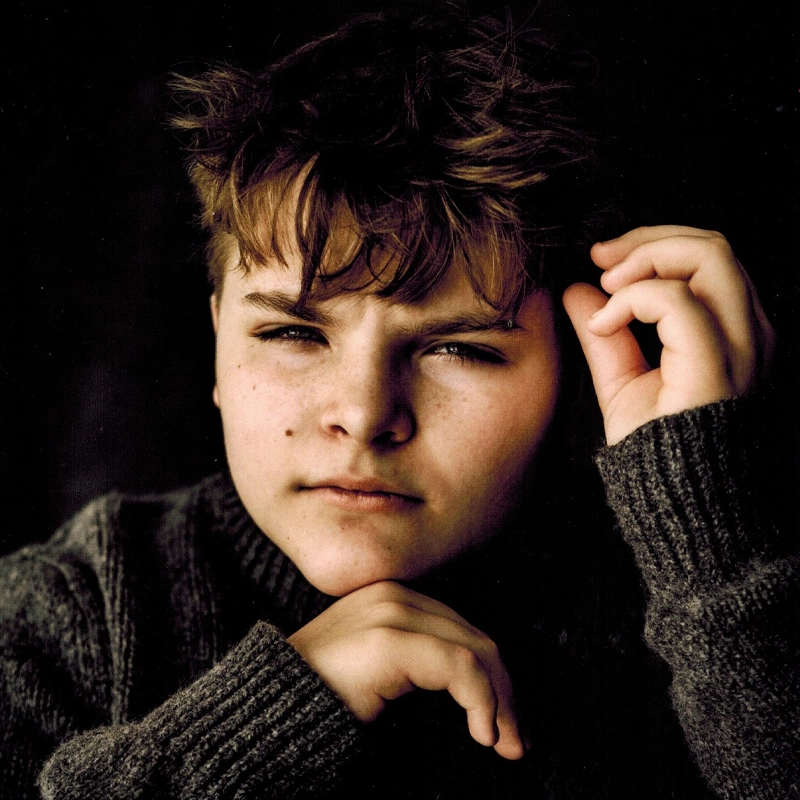 A young person with tousled brown hair and fair skin wears a chunky, dark knit sweater. They look intently at the camera with a neutral expression, one hand raised by their face and the other resting near their chin. The background is dark, drawing all attention to Colton Piontkowski’s contemplative gaze.