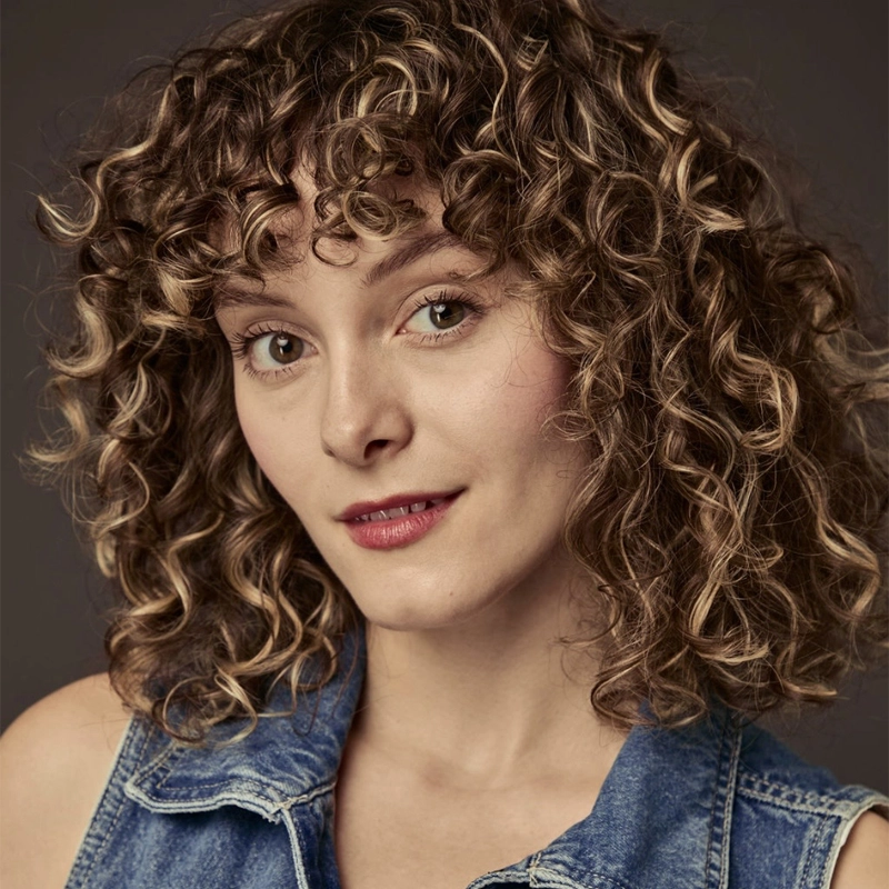A young woman with light skin and chin-length, blonde-brown tightly curled hair with bangs looks at the camera. She has hazel eyes, light makeup, and a subtle smile. She is wearing a sleeveless blue denim top. The background is plain and dark, drawing focus to Grace Pompei’s thoughtful expression.