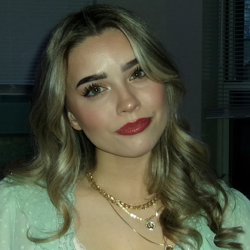 A young woman with wavy, light brown hair and bold makeup wears layered gold necklaces and a light green top. She looks at the camera with a slight smile, reminiscent of Haley Raffaele’s effortless style. The background includes a window with blinds and dim indoor lighting.