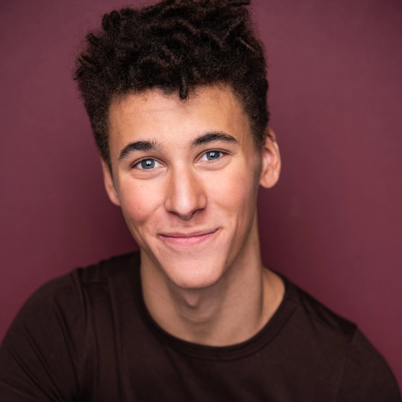 Zakari Emanuel Ramos, a young man with short curly dark hair and blue eyes, smiles softly. He is wearing a plain dark shirt and is posed in front of a solid maroon background, looking directly at the camera.