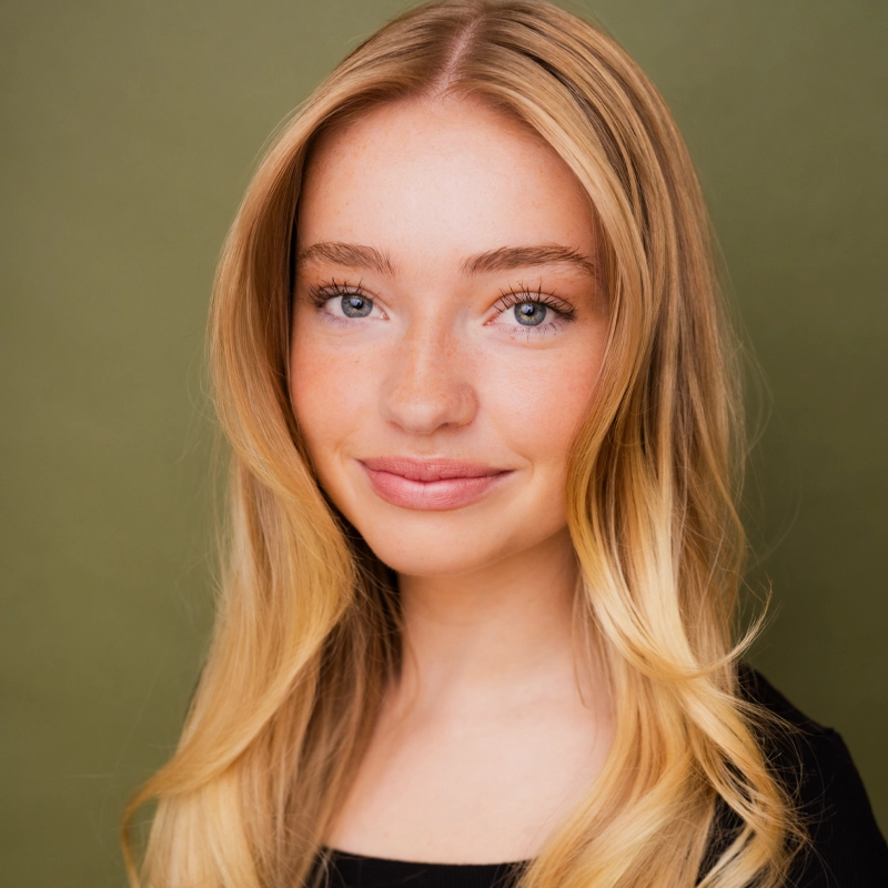 A young woman with long, wavy blonde hair smiles softly. She has fair skin, blue eyes, and light freckles. She wears a black top and is posed in front of a plain olive-green background.