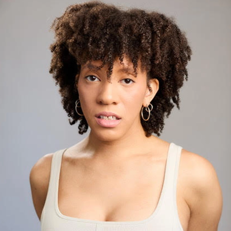 A professional headshot of Jayae Riley Jr., a Black woman with dark, curly, shoulder-length hair and small gold hoop earrings. She is wearing a light-colored tank top and looking directly at the camera with a neutral, thoughtful expression against a soft, light gray studio background.