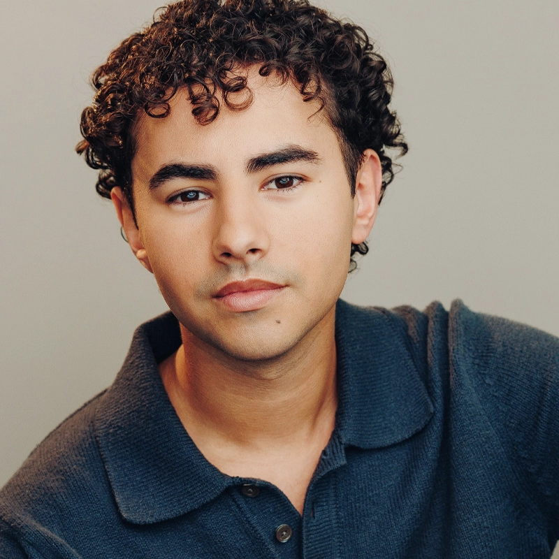 A young person with short, curly dark hair and light skin looks directly at the camera, wearing a dark blue collared shirt. The background is plain and softly lit, highlighting Adrian Rivera’s calm and confident expression.