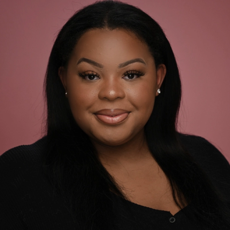 A woman with straight black hair, subtle makeup, and stud earrings smiles gently in front of a plain pink background. She wears a black top and looks directly at the camera, exuding confidence and warmth.