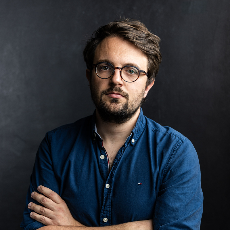 A man with short brown hair, beard, and round glasses stands against a dark background. He wears a blue button-up shirt with sleeves rolled, arms crossed, and looks directly at the camera with a neutral expression. The lighting is soft and moody.