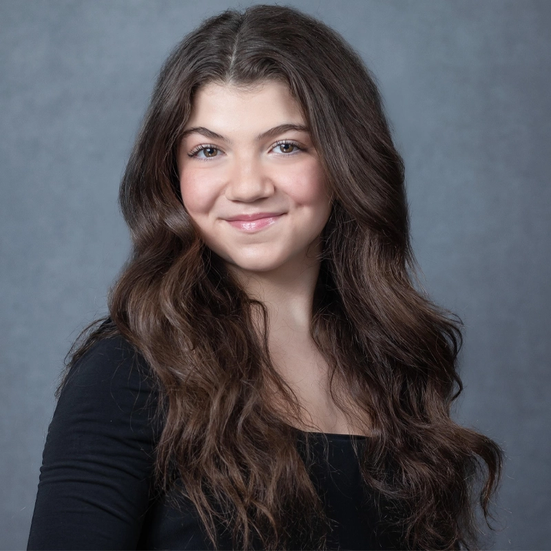 A young woman with long, wavy brown hair and fair skin smiles at the camera. Adabelle Ross wears a black top and stands against a plain, light gray backdrop. Her expression is warm and friendly.