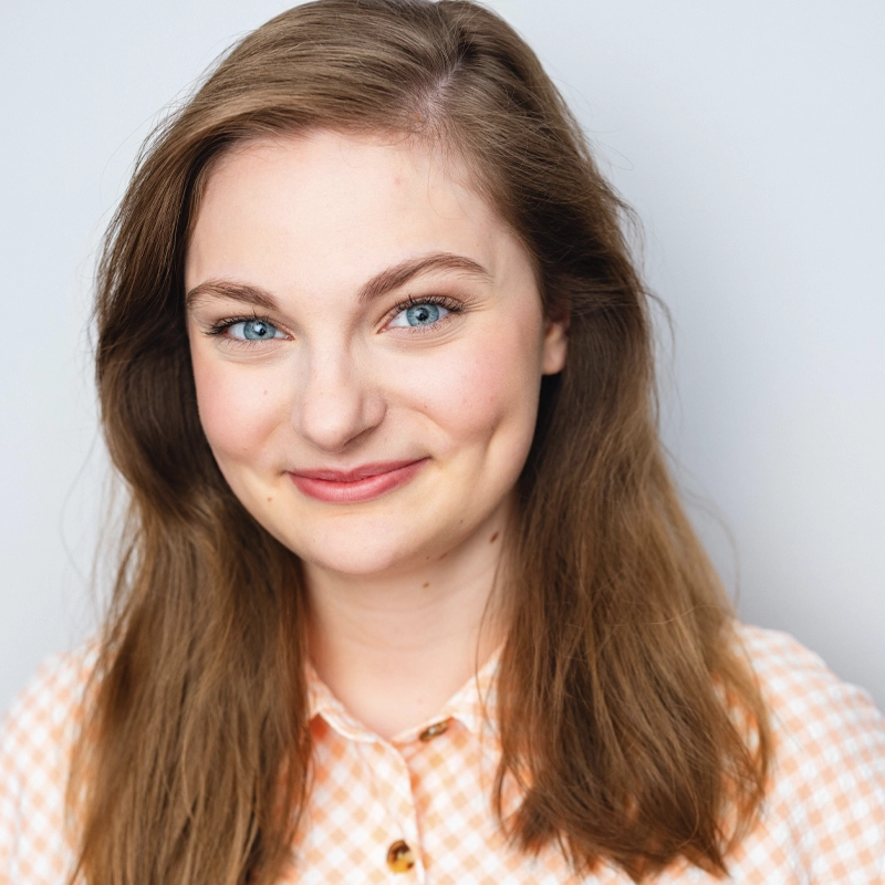 A young woman with long, light brown hair and blue eyes smiles slightly at the camera. Annika Rudolph wears a peach and white checkered shirt, and the background is plain light gray. The image is well-lit, highlighting her clear skin and natural look.
