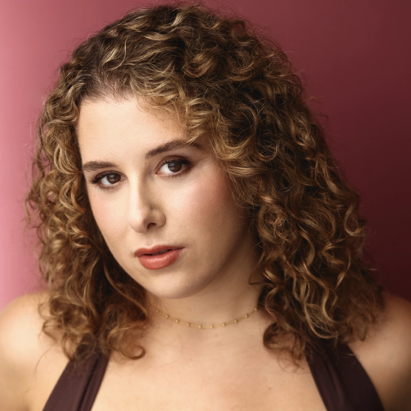 A woman with shoulder-length, curly blonde hair poses against a rose-pink background. She wears a sleeveless brown top and a delicate gold necklace. Her makeup is subtle, with natural tones on her lips and eyes, and she gazes confidently at the camera.