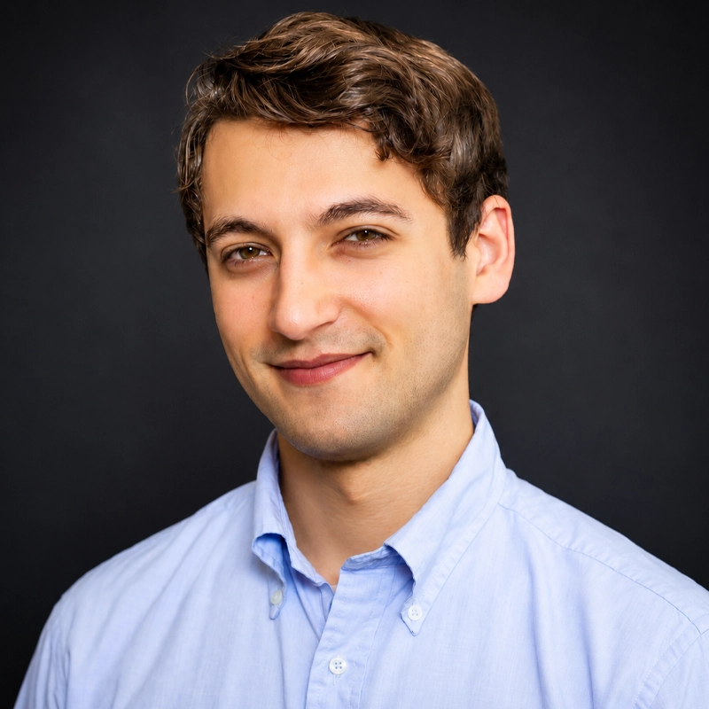 A young man with short, wavy brown hair and fair skin smiles softly. He wears a light blue, button-down shirt. The background is plain and dark, highlighting his face and upper torso. The lighting is even and professional, suggesting a formal portrait.