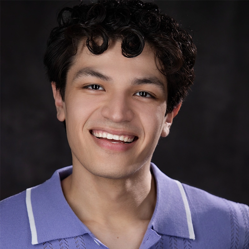 A young person with curly dark hair smiles at the camera. They wear a light purple collared shirt with white trim. The background is dark and blurred, focusing attention on their face. The image conveys a warm and friendly expression, capturing the welcoming presence of Salvador Diaz-DeBose.