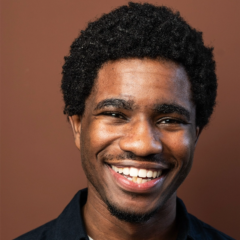 A young man with short curly hair smiles warmly. He has dark skin, a trimmed mustache and beard, and wears a dark shirt. The plain brown background keeps the focus on his happy expression.
