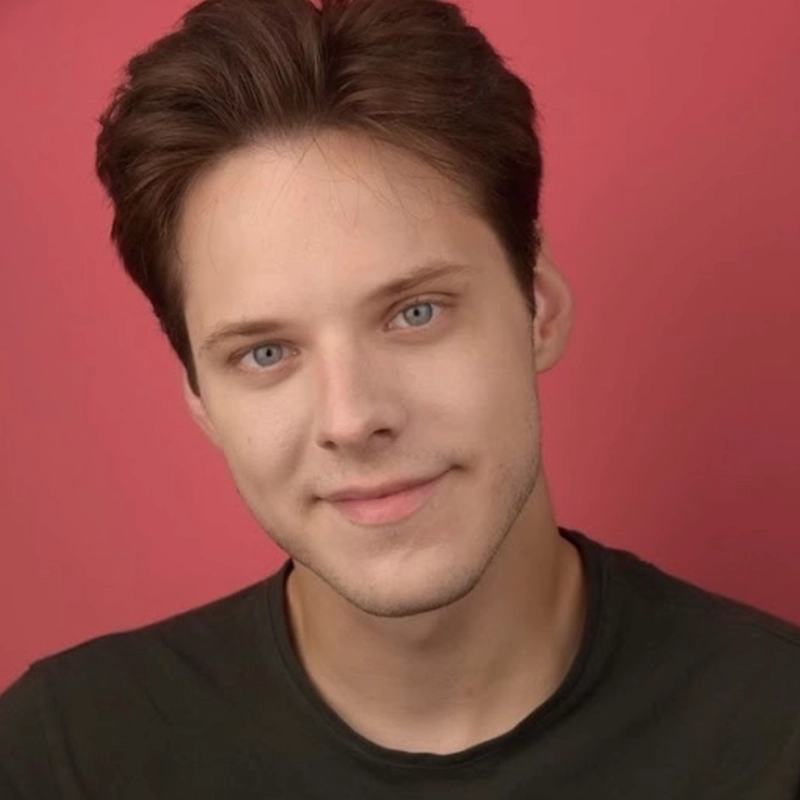 A young person with short, brown hair and light skin smiles gently at the camera. They have blue eyes and wear a dark, crew-neck shirt. The background is a solid, bright pink color, lending a vibrant backdrop to Kendall Tubbs’ friendly demeanor.