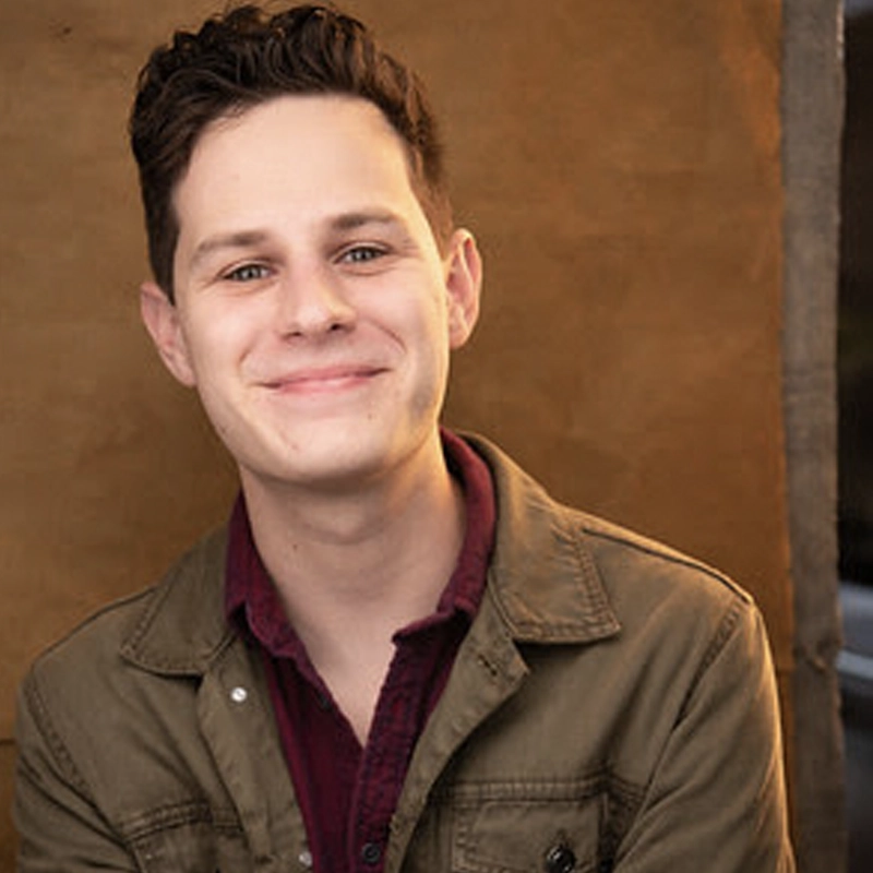 A young person with short, dark brown hair smiles warmly. Jimmy Schumacher wears a maroon collared shirt under an olive green jacket. The background is a solid, textured tan surface, softly lit, giving the photo a casual, friendly atmosphere.