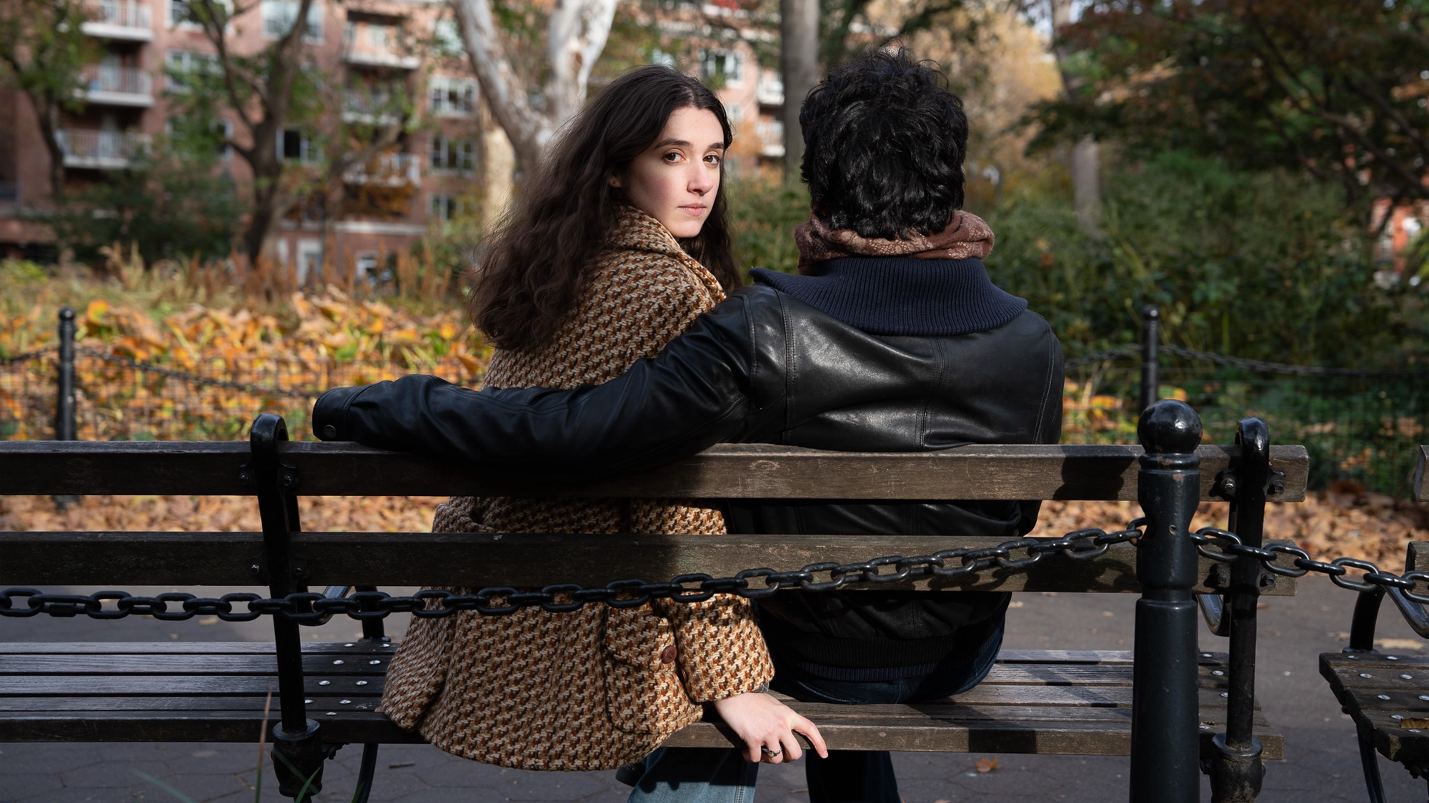 A young woman and a man sit on a park bench. The woman, in a brown houndstooth coat, looks over her shoulder at the camera; the man wears a black jacket with his arm around her. Autumn trees and buildings are visible in the background, setting an intimate scene reminiscent of The Maker & The Muse, captured in quiet elegance by Schuyler Iona Press.