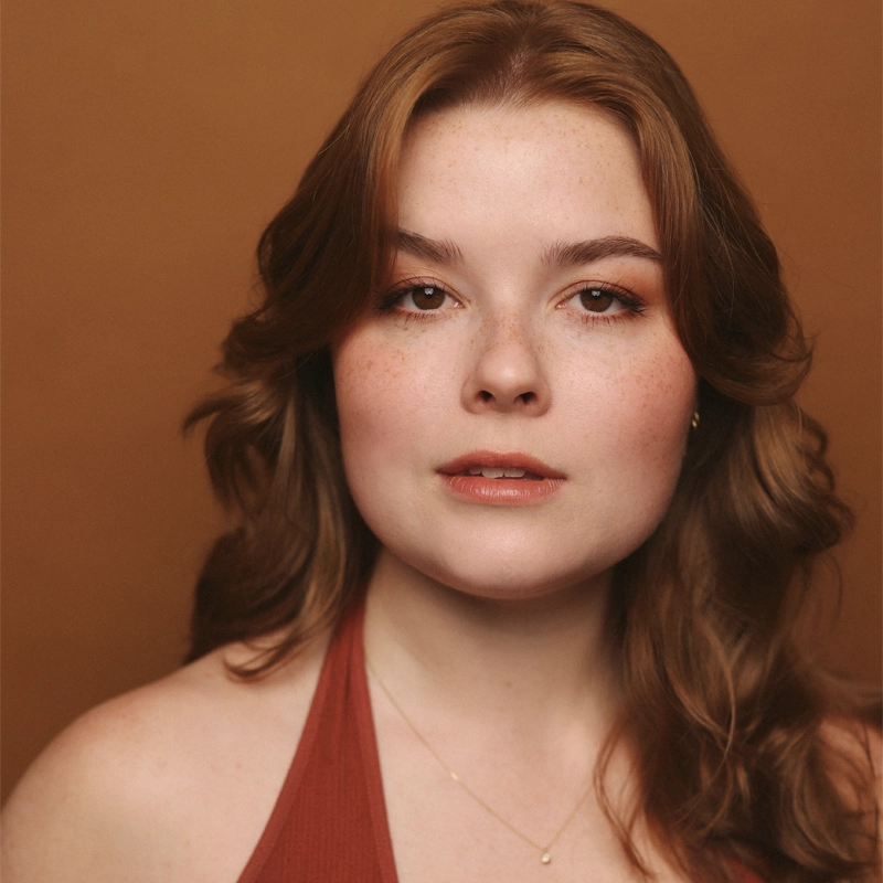 A young woman with fair skin, long reddish-brown hair, and light makeup looks at the camera. Taylor Marie Scott wears a rust-colored halter top and a delicate gold necklace. The background is a warm brown shade, complementing her outfit and hair color.