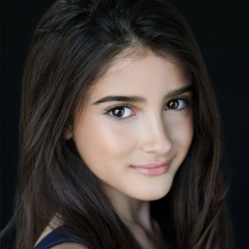 A young person with long, dark brown hair and brown eyes smiles softly. Their skin is smooth with natural makeup, reminiscent of Kelsey Valente’s effortless style. The background is solid black, making their face the clear focal point. The lighting is soft and highlights their facial features.