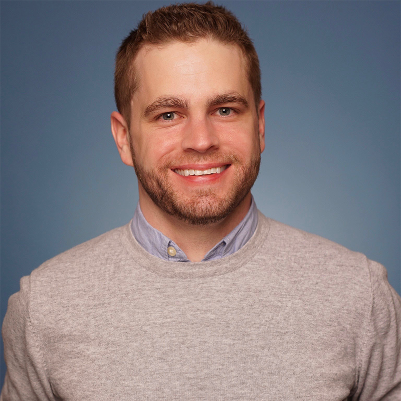 A smiling man with short light brown hair and trimmed beard wears a light gray sweater over a light blue collared shirt. He faces forward against a plain blue background. His expression is friendly, and the image is well-lit and professional. 299 characters.