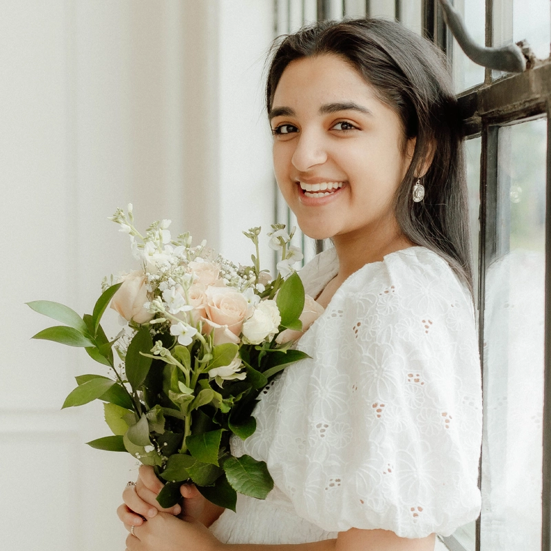 Sasha Shetty, a young woman with long dark hair, stands by a window smiling gently, dressed in a white eyelet dress and holding a bouquet of pale roses and greenery. Pearl earrings adorn her ears as she basks in the softly lit, elegant setting.