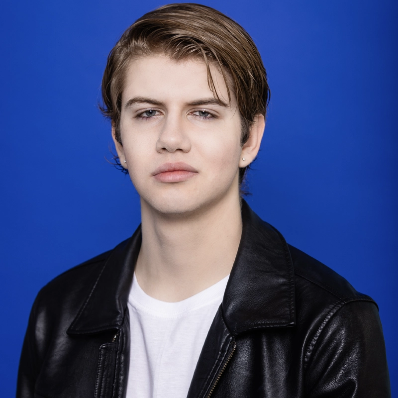 A young person with fair skin, straight light brown hair swept to the side, and light eyes looks at the camera. They wear a black leather jacket over a white T-shirt. The background is bright blue.