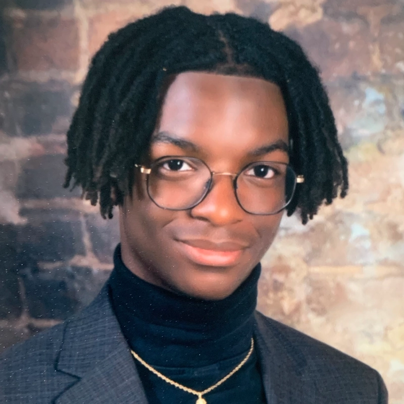 A young person with medium-length dreadlocks, wearing round glasses, a dark turtleneck, a grey blazer, and a gold necklace, poses against a blurred brick wall background. Justin Taylor Smith looks directly at the camera with a slight smile.