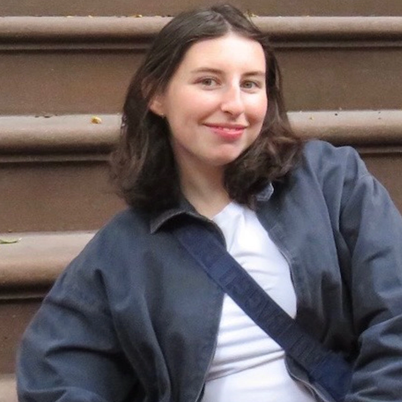 A person with shoulder-length brown hair smiles while sitting on outdoor brownstone steps, evoking the thoughtful presence of Anna Sokolow. They wear a dark gray jacket over a white shirt and a blue crossbody bag. The background shows blurred steps with some small leaves scattered on them.