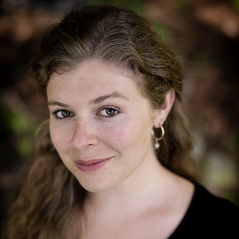 A young woman with wavy light brown hair, wearing hoop earrings and a black top, smiles softly at the camera. The background is blurred with earthy tones, highlighting Allison Spann’s face.