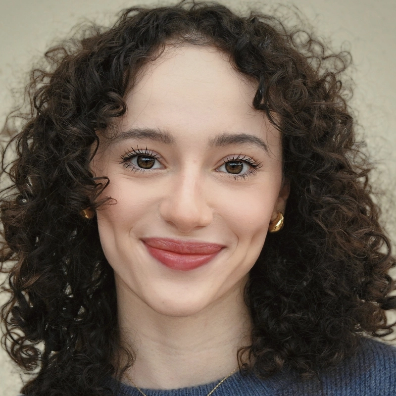 Gianna Spironello, a young woman with fair skin and curly brown hair, smiles warmly. She wears gold hoop earrings, a gold necklace, and a blue top. The background is neutral and out of focus, placing attention on her expressive eyes and natural makeup.