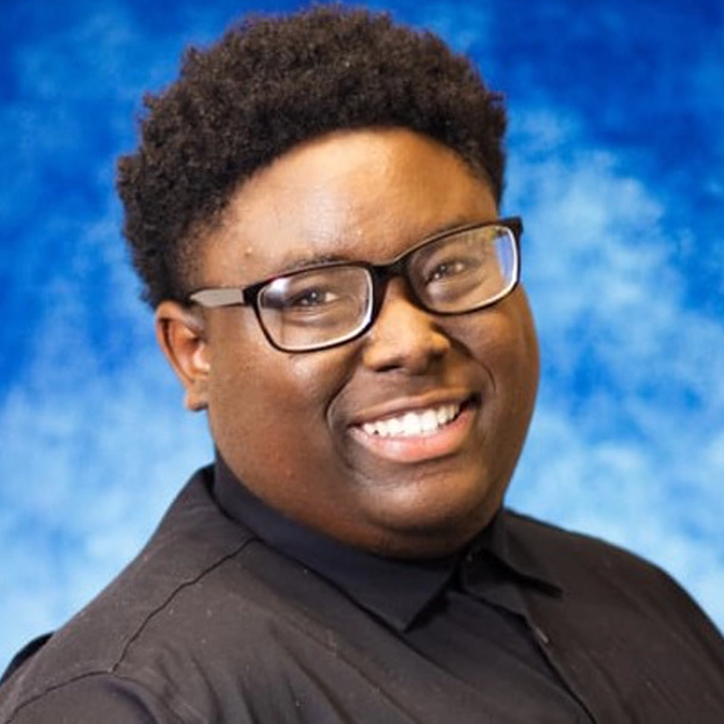 A smiling person with short curly hair and rectangular glasses wears a black collared shirt. The background is a soft blue with lighter areas, creating a gentle, professional effect. Daniel Stanley is facing the camera with a friendly expression.
