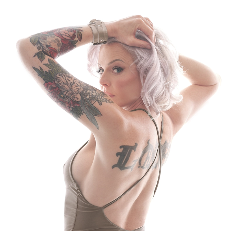 A woman with pale skin and platinum blonde hair poses with her arms raised, showing detailed floral tattoos on her left arm and “LOVE” tattooed across her upper back. She wears a strappy taupe top and a studded bracelet, standing against a bright white background.