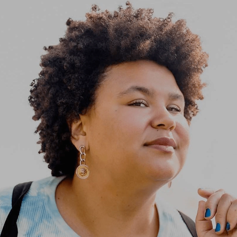 Regina Strayhorn, with short, curly hair and gold earrings, gazes confidently to the side. She wears a light tie-dye shirt and has blue nail polish. The background is light and out of focus, highlighting her calm and self-assured expression.