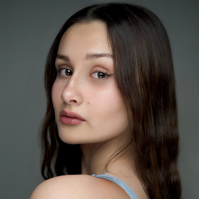 A young woman with long brown hair, clear fair skin, and brown eyes looks over her shoulder toward the camera. Grace Suarez has natural makeup, full lips, and is wearing a light blue sleeveless top. The background is a plain gray.