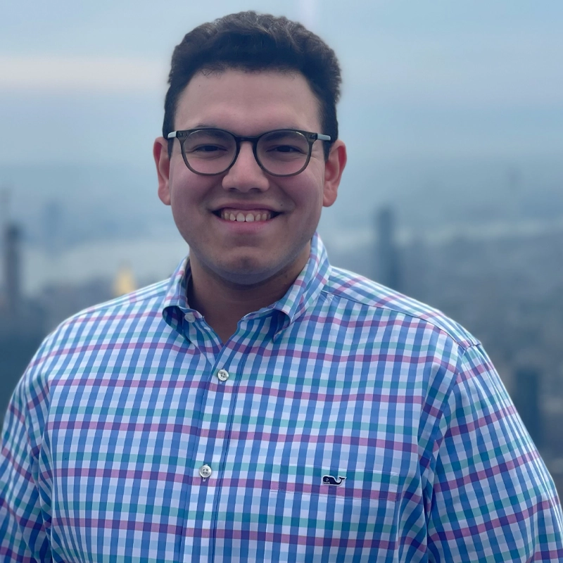 A smiling man with dark hair and glasses, Matthew Sullivan, wears a colorful plaid button-down shirt, standing outdoors with a blurred city skyline in the background on a cloudy day.