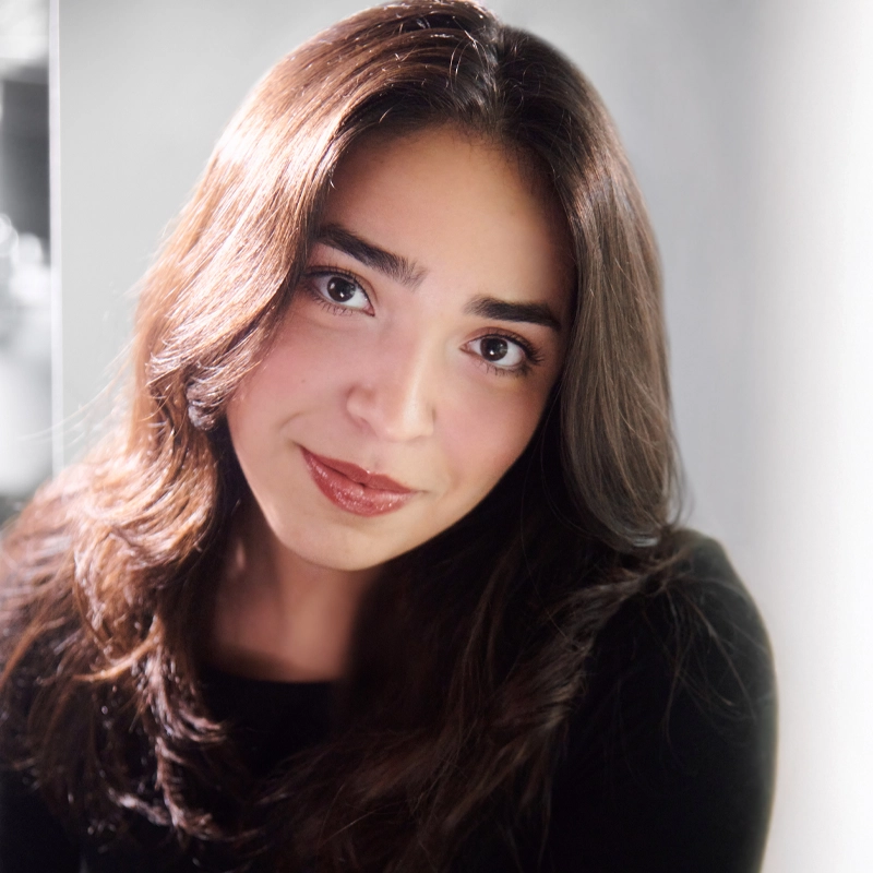 A woman with long, wavy brown hair, wearing a black top, smiles softly at the camera. She has light skin, brown eyes, and natural makeup. The background is softly blurred, drawing focus to her face. Light shines on her hair, adding warmth.