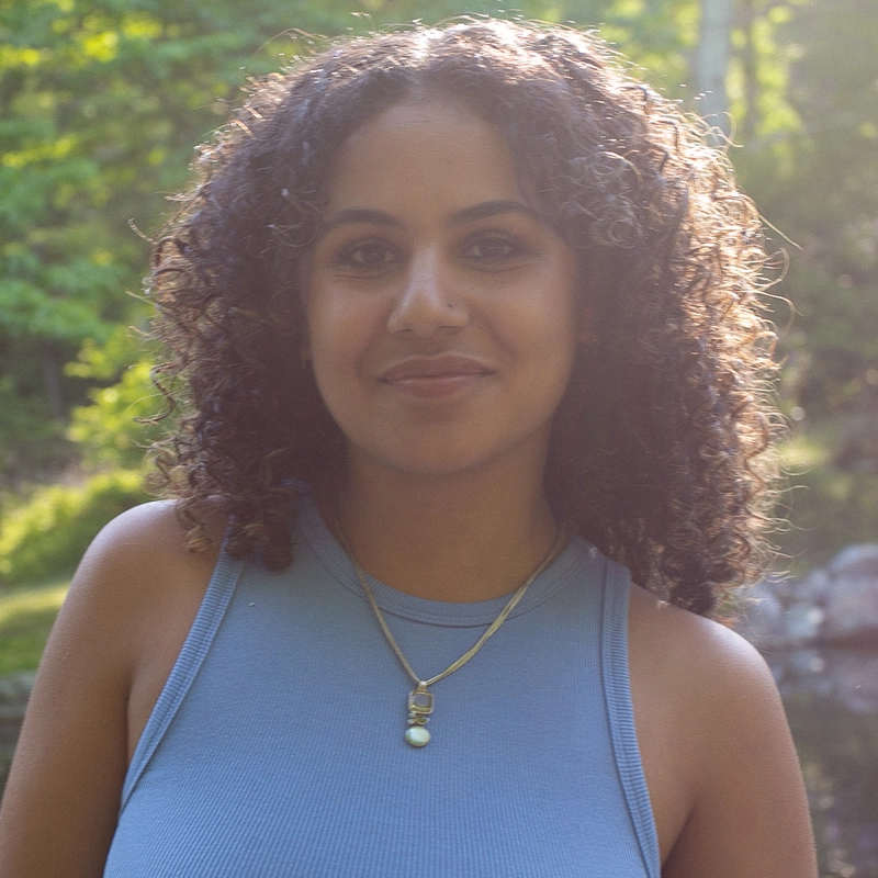 Lula Talenfeld, a woman with curly hair, smiles outdoors, wearing a light blue sleeveless top and two gold necklaces. Green trees and sunlight fill the blurred background, suggesting a park or natural setting.