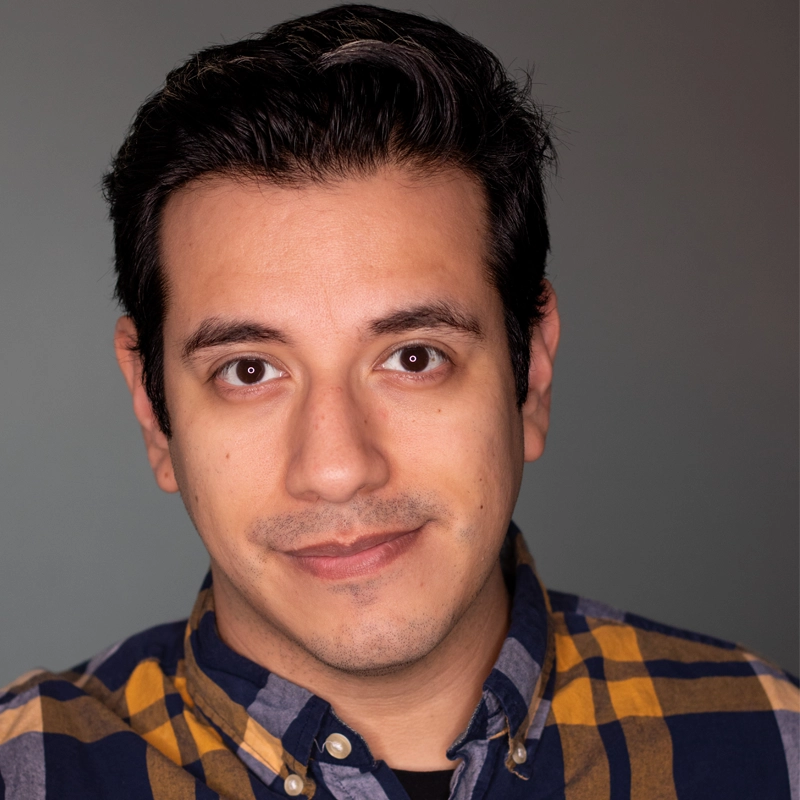 A man with short dark hair and brown eyes looks at the camera with a slight smile. He wears a yellow and blue plaid shirt over a black T-shirt. The background is plain and softly lit, creating a warm, professional portrait atmosphere—an image that could easily feature Jeffrey Quintana Tapia.