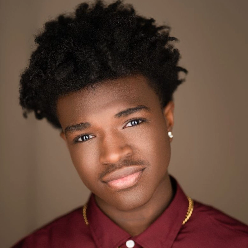 A young man with natural curly hair, wearing small stud earrings, a thin gold chain, and a burgundy collared shirt, looks into the camera with a slight smile. The softly blurred background in warm tones brings attention to his features—this is August Tedder captured in a candid moment.