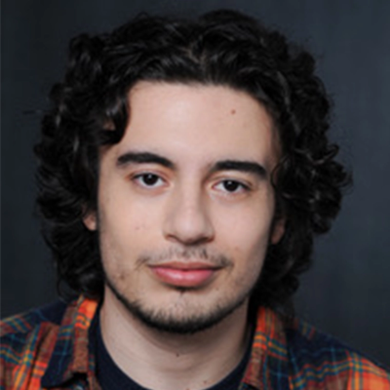 A young man with shoulder-length curly dark hair and light facial hair. He is wearing a plaid shirt and looking directly at the camera.