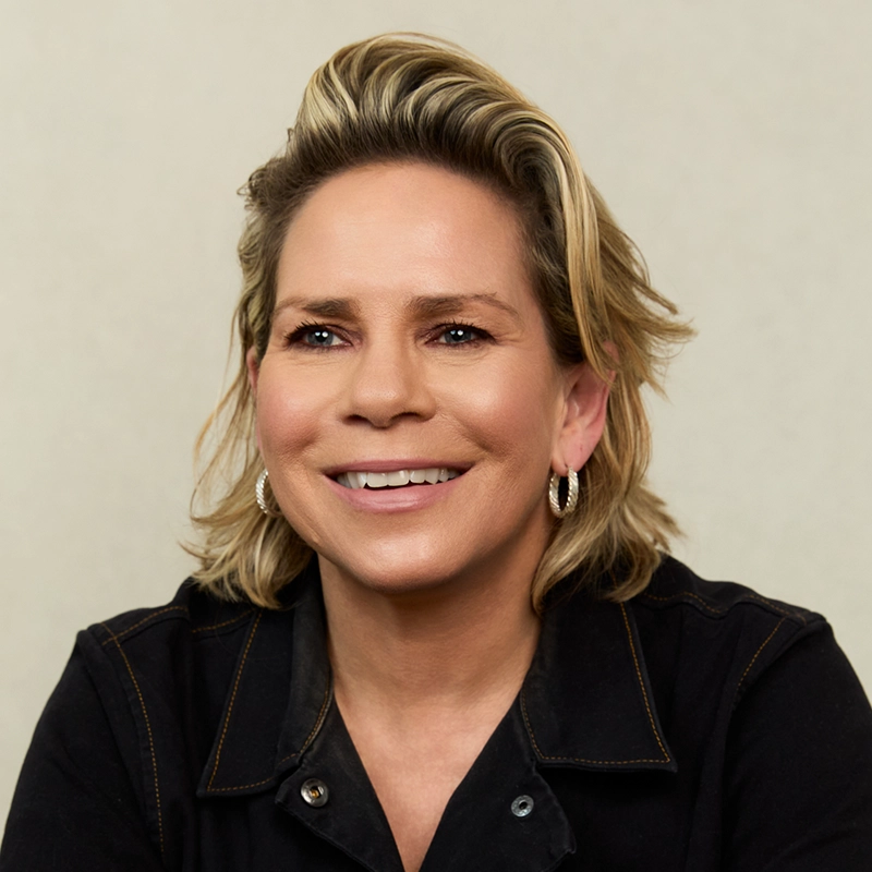 A smiling person with short, tousled blonde hair and hoop earrings, wearing a black collared shirt, sits in front of a plain light background—capturing the confident presence of Tracy E. Gilchrist.