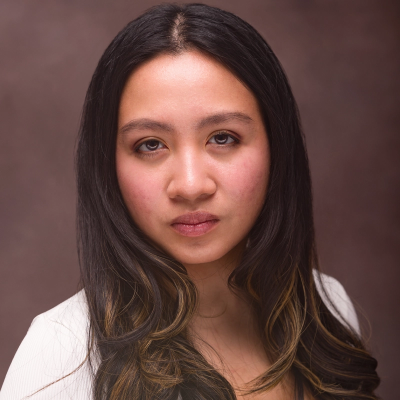 A young woman with long, wavy dark hair and subtle highlights stares confidently at the camera. She has a neutral expression, light makeup, and is wearing a white top. The background is softly blurred in shades of brown.