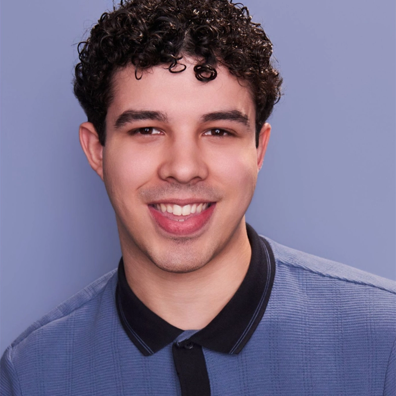 A young man with short curly hair smiles at the camera. He is wearing a blue collared shirt and is posed against a plain light purple background. The lighting is even, highlighting his cheerful expression.