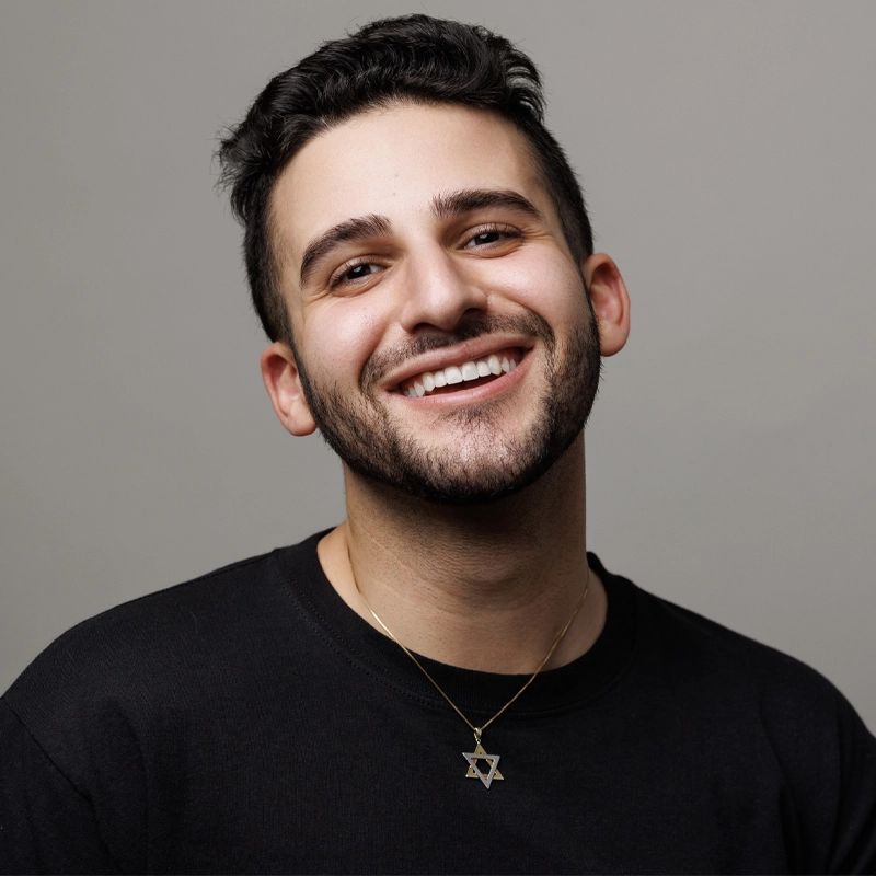 A young man with short dark hair and a trimmed beard smiles warmly at the camera. He wears a black shirt and a gold necklace featuring a Star of David pendant. The background is plain and light gray, giving Michael Valdes an approachable, modern presence.