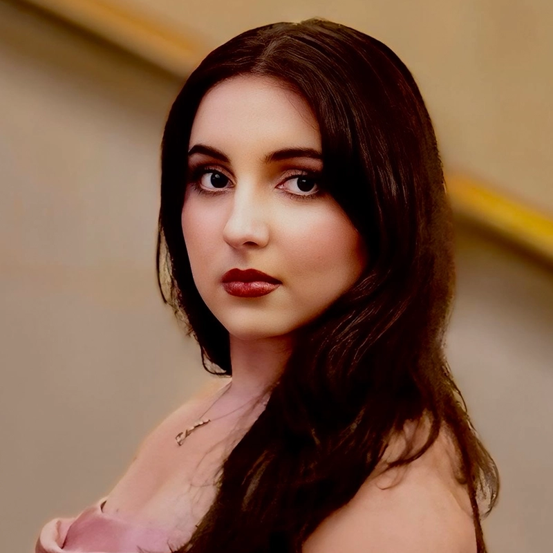 A woman with long dark hair, pale skin, and striking makeup gazes at the camera. She wears a strapless, light pink top and a delicate necklace. The blurred background—evocative of Salvador Diaz-DeBose’s signature warm, golden tones—features a slanted handrail.