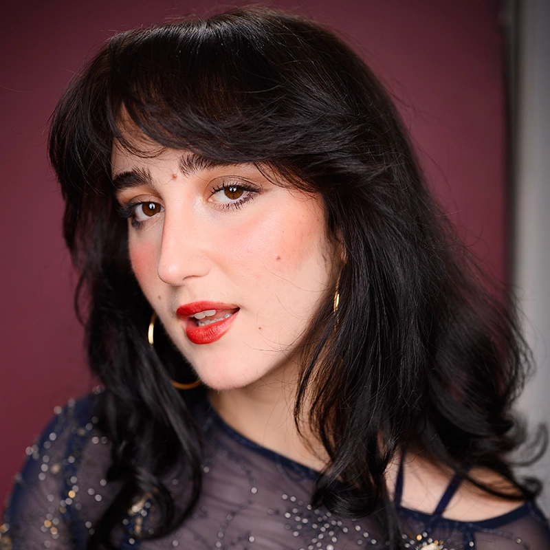 A person with long black hair, wearing gold hoop earrings and a sheer, sparkly navy top, poses against a mauve background. Cooper Brown has light skin, bold eyebrows, subtle blush, and red lipstick, with a soft, confident expression.