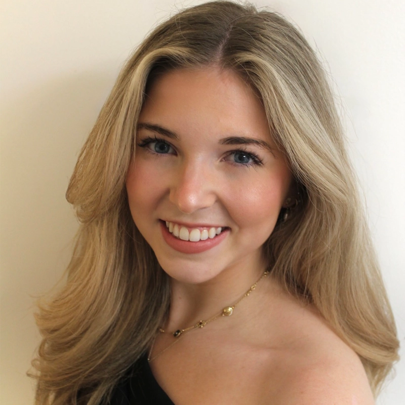 A young woman with long, wavy blonde hair smiles at the camera. She has blue eyes, light skin, and wears a delicate gold necklace and a black off-the-shoulder top. The background is plain and light-colored, highlighting the graceful presence of Mary Ashton VanFossen.